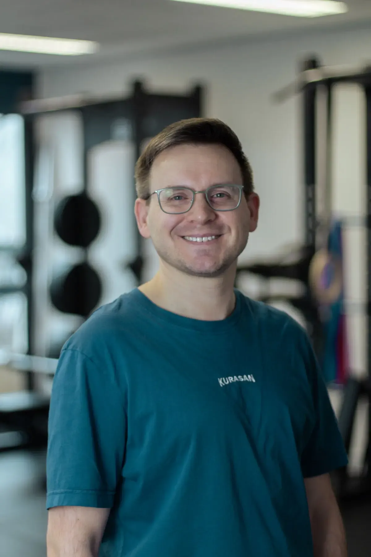 Lächelnder Mann mit Brille in Sportshirt, Porträt im Fitnessstudio mit Trainingsgeräten im Hintergrund.