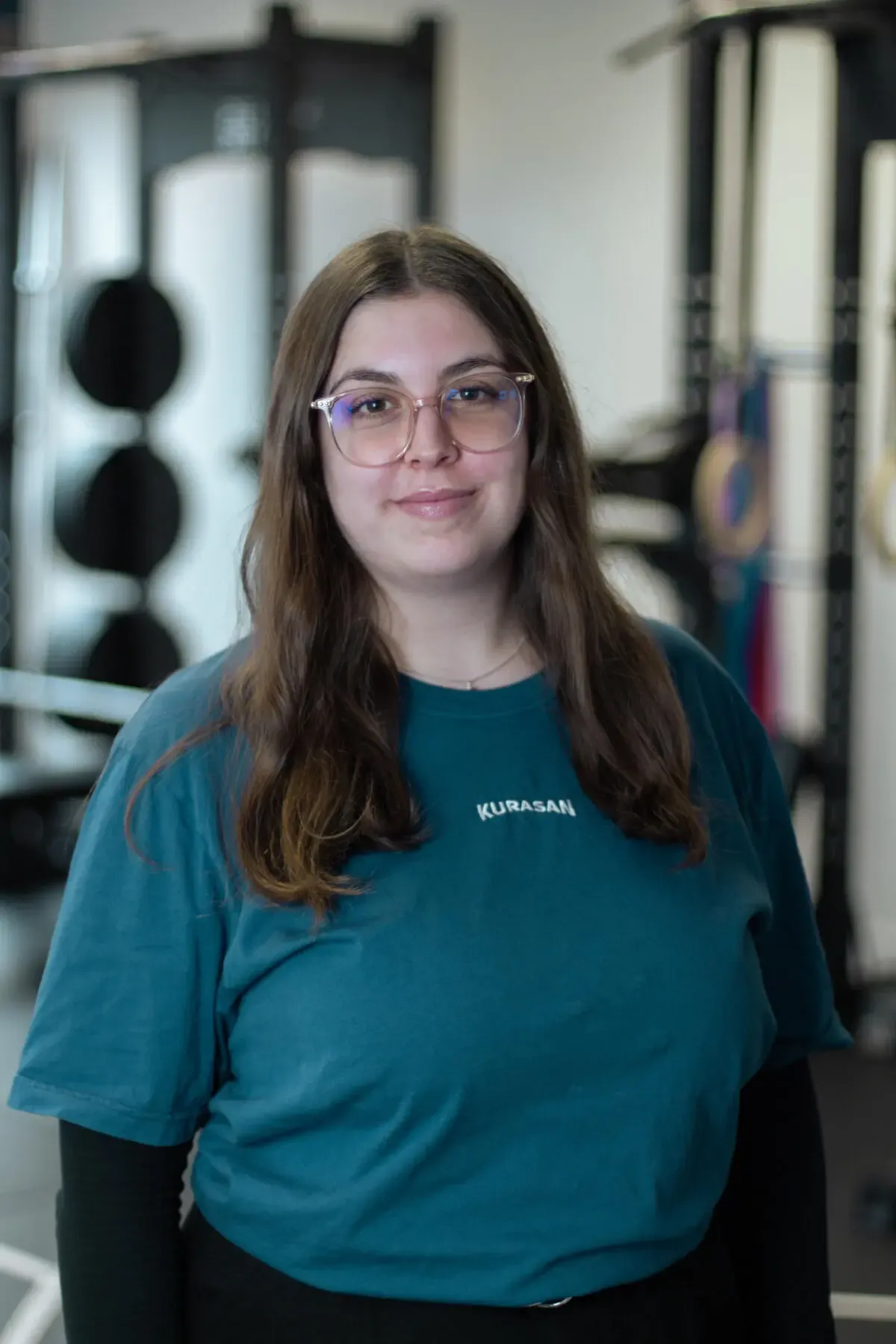 Porträt einer Trainerin mit Brille im Fitnessstudio, im Hintergrund Trainingsgeräte und Gewichte.