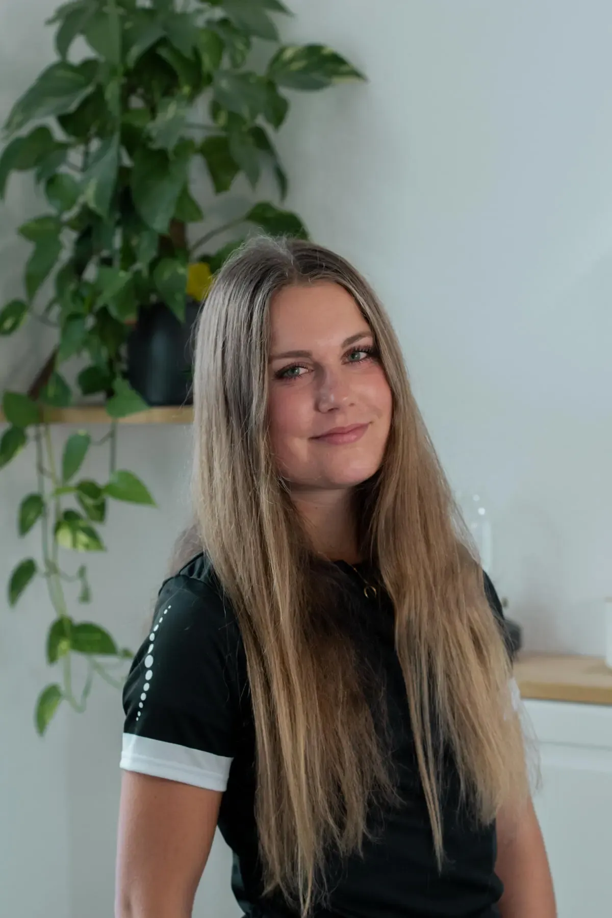 Portrait of a smiling woman with long hair indoors, with a leafy houseplant on a shelf behind her.