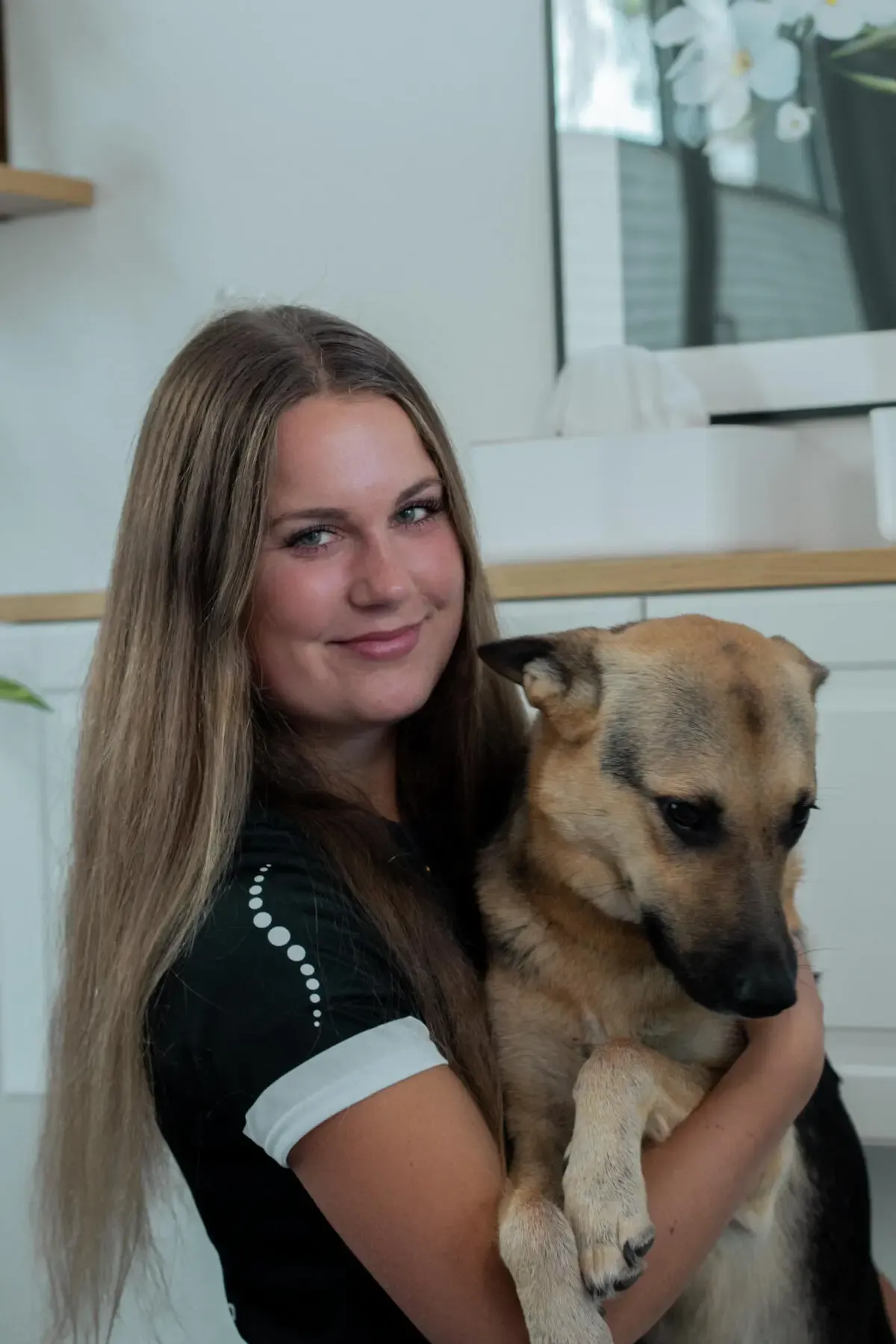 Smiling woman with long hair holding a tan dog in a bright indoor room.
